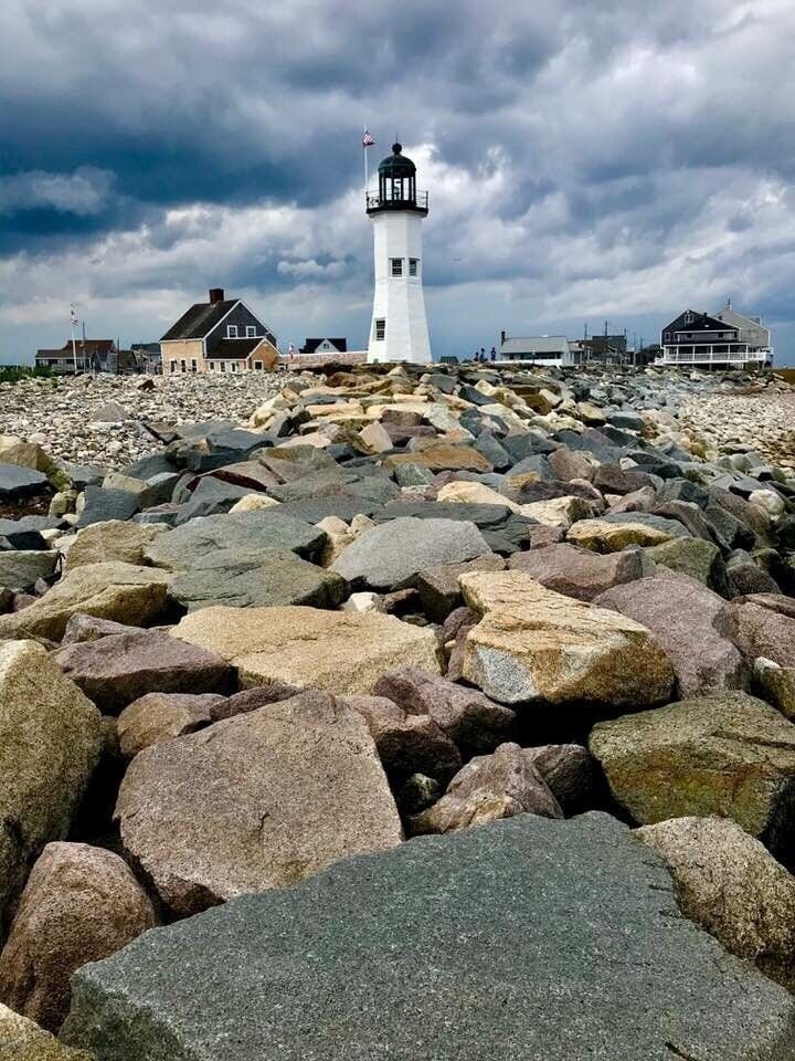 A lighthouse on a rocky shore with buildings in the background.