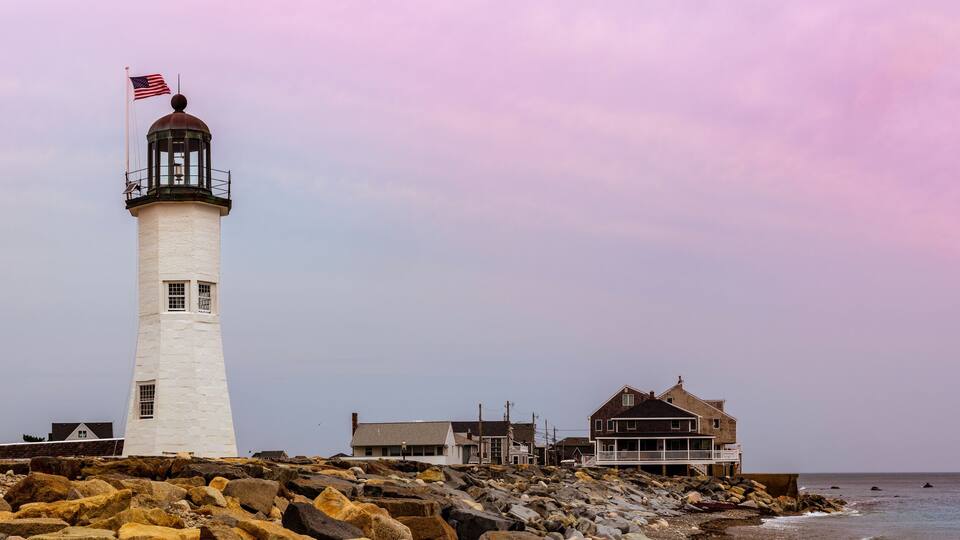 A lighthouse with an American flag, surrounded by rocky shoreline and coastal buildings.