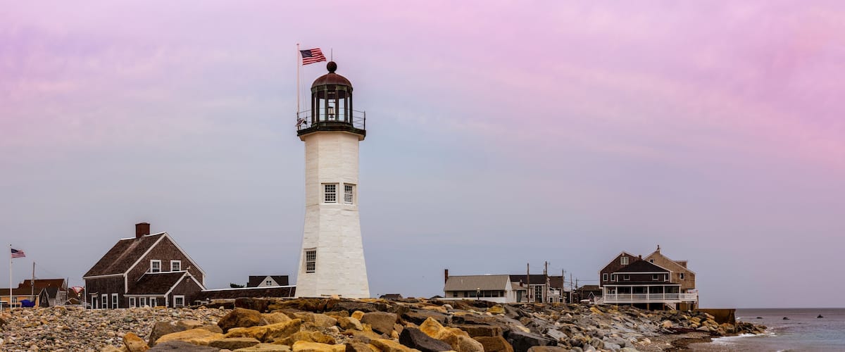 A lighthouse with an American flag, surrounded by rocky shoreline and coastal buildings.