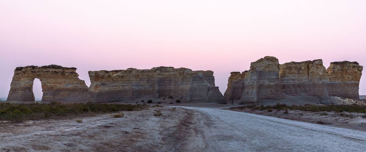A dirt road leading to a series of rock formations with a pink sky in the background.