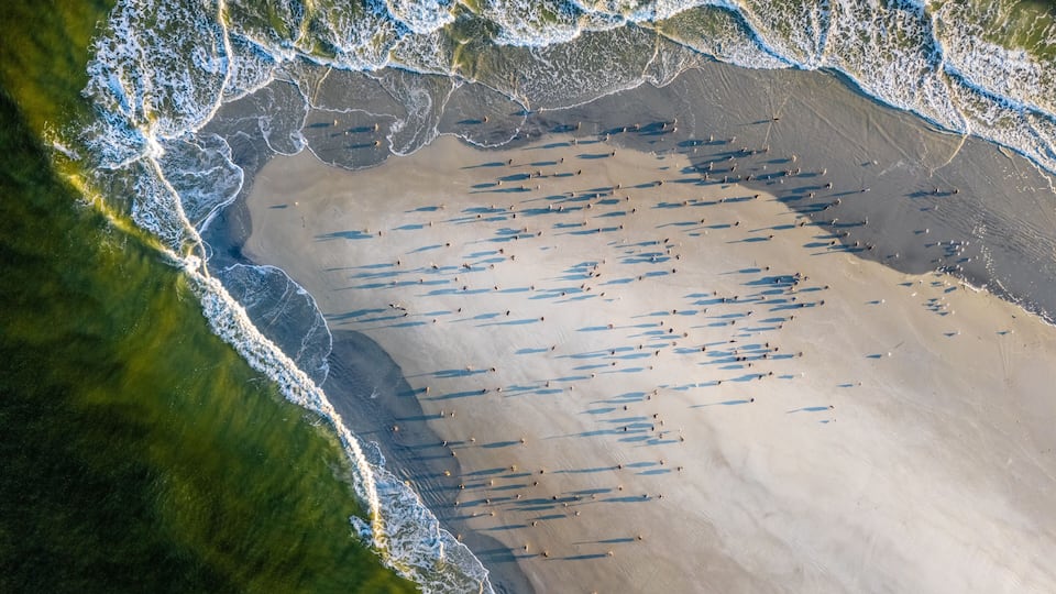 An aerial view of a beach with waves, sand, and sparse vegetation.