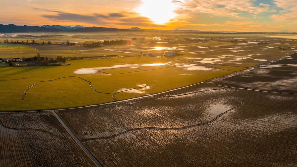 A vast agricultural landscape with fields, a river, and distant mountains under a sunset sky.