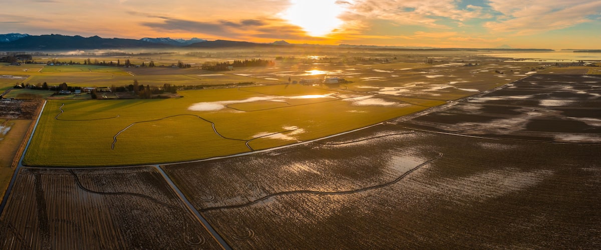 A vast agricultural landscape with fields, a river, and distant mountains under a sunset sky.