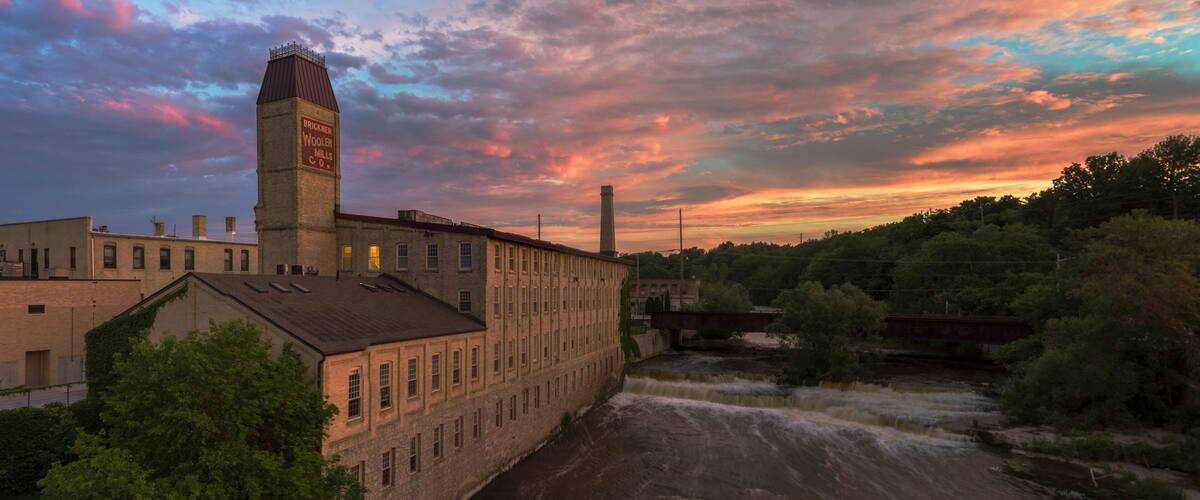 A historic industrial building with a prominent tower, a river flowing through the scene, and a bridge in the background.