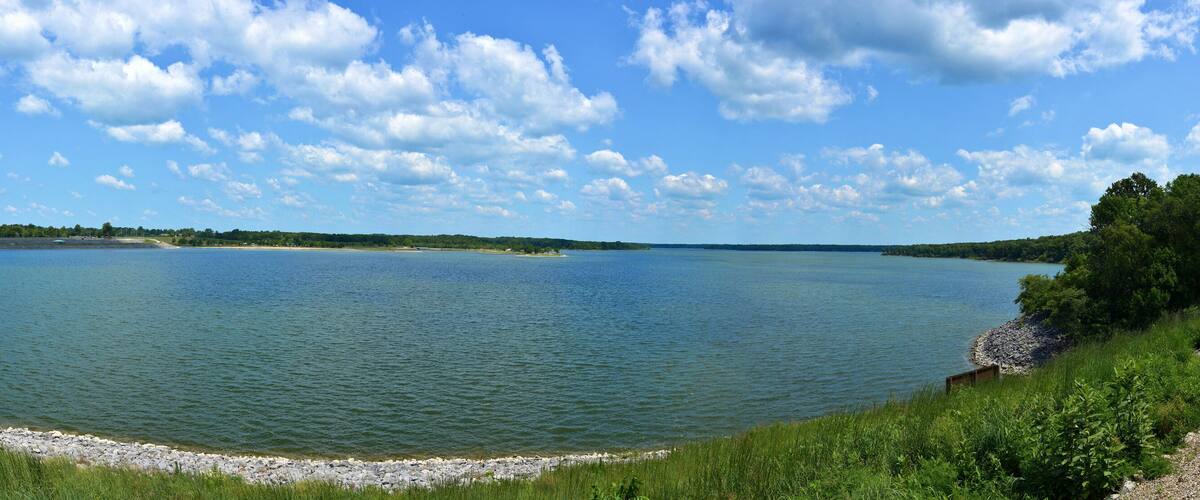 A large lake with a rocky shoreline, surrounded by greenery and trees under a partly cloudy sky.