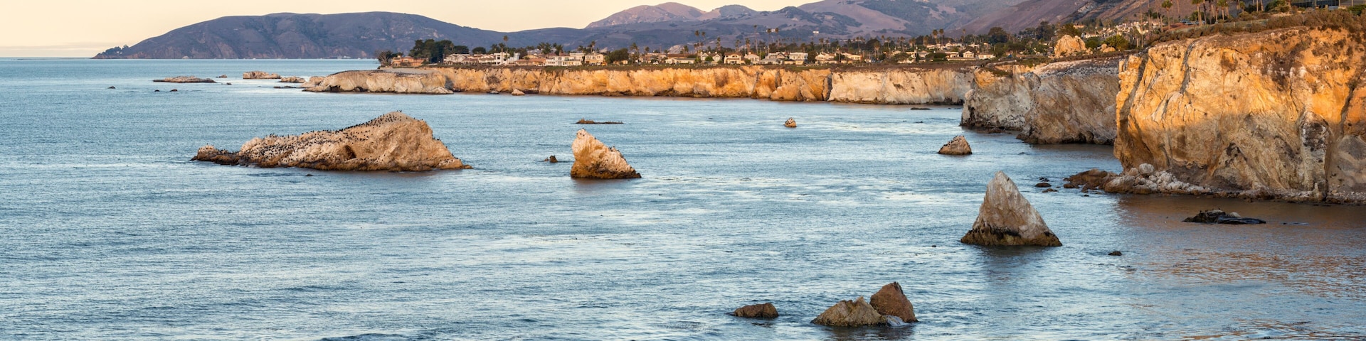 A coastal landscape with rocky outcrops in the water, a distant mountain range, and a clear sky.