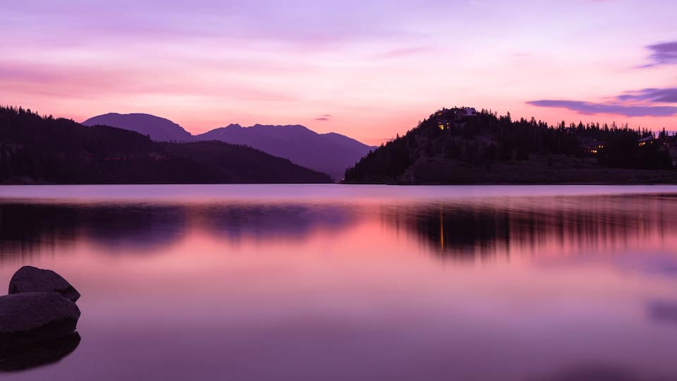 A calm lake at dusk with mountains in the background and a forested hill on the right.