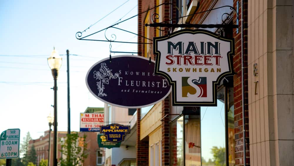 A street sign for Main Street in Kowhegan, a sign for a florist, and a parking sign.