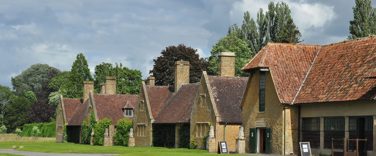 A row of traditional stone houses with red-tiled roofs, surrounded by lush green grass and trees.