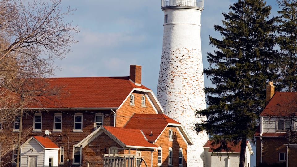 A lighthouse with a red roof, a brick building with a white trim, and a large evergreen tree.