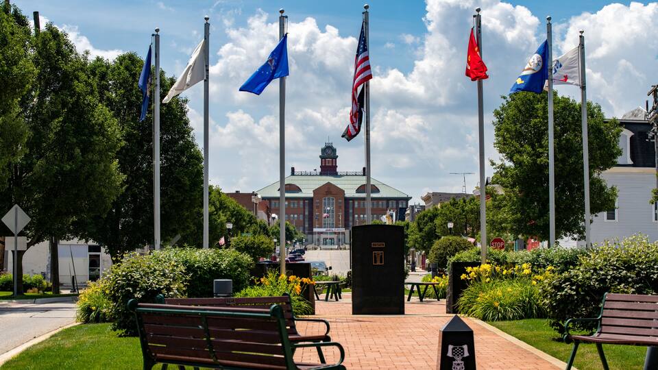A flag display with multiple flags, a building in the background, and benches in a park.