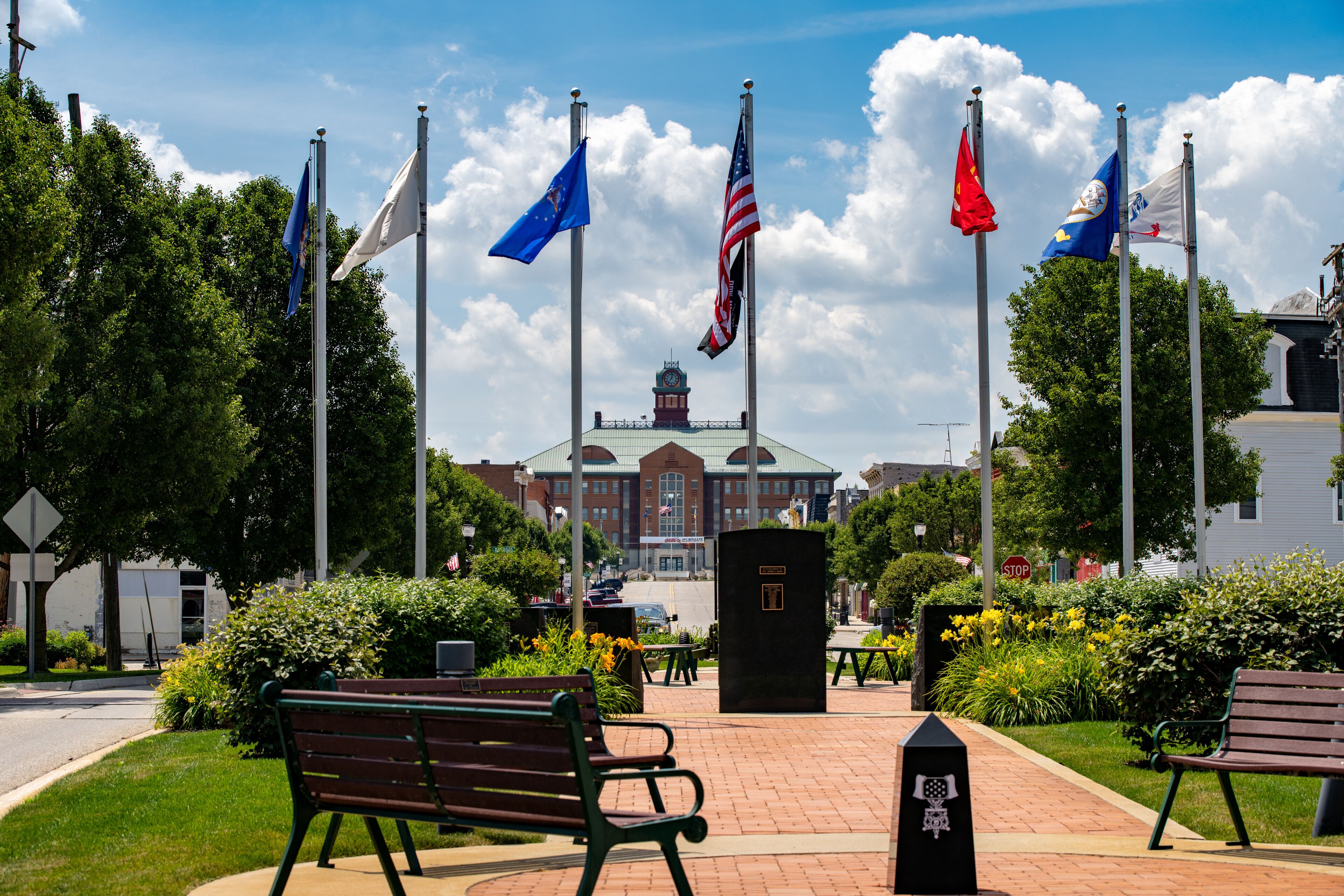A flag display with multiple flags, a building in the background, and benches in a park.