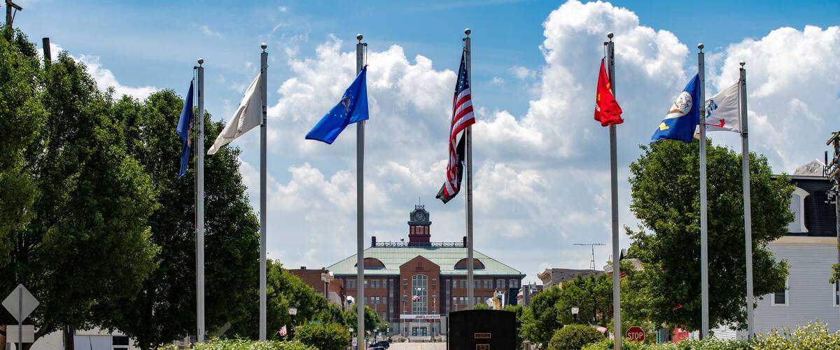 A flag display with multiple flags, a building in the background, and benches in a park.