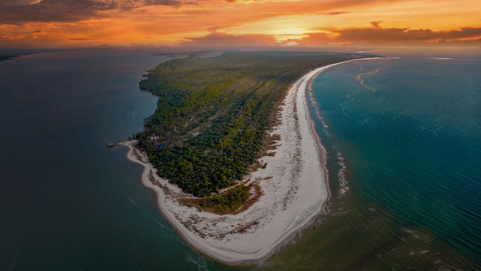 An aerial view of a coastal landscape with a narrow strip of land, dense vegetation, and a curving coastline.