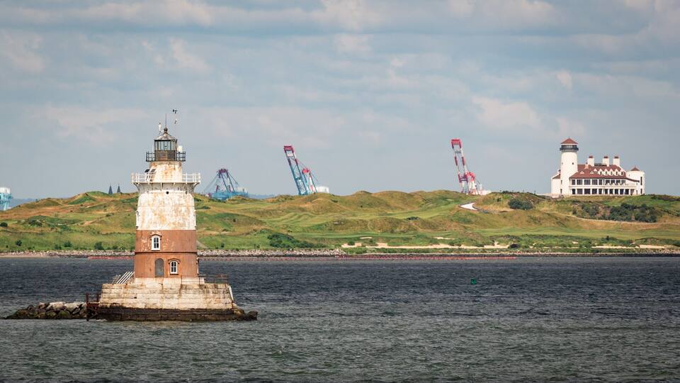 A lighthouse, a large white building, and several cranes on a hilly landscape by the water.