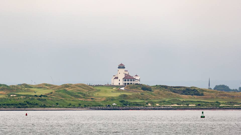 A coastal landscape with a lighthouse on a hill, a golf course, and a calm sea.