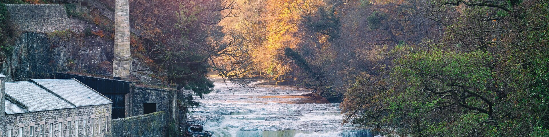 A river flowing through a forested area with a stone building on the left.