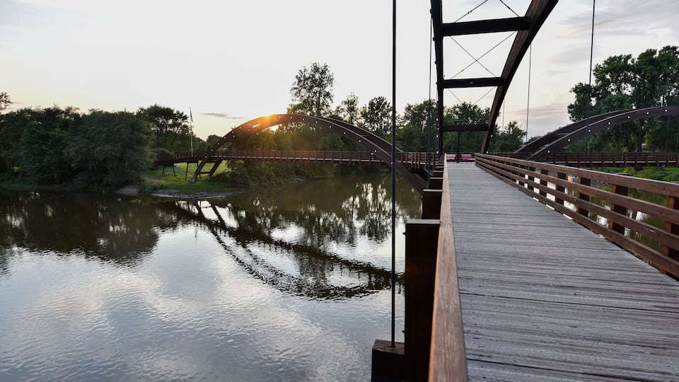 A wooden bridge over a calm river with trees on either side and the sun setting in the background.