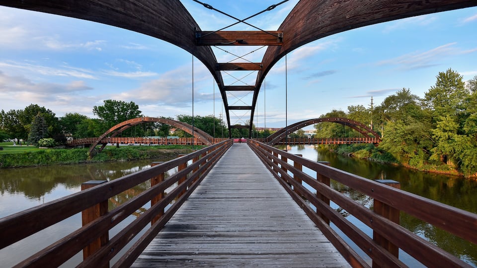 A wooden pedestrian bridge over a calm river, surrounded by lush greenery and trees.