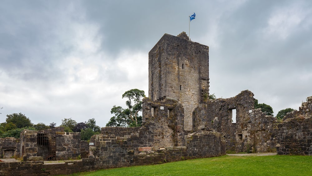Une tour de pierre historique, surmontée d’un drapeau, entourée de ruines et de verdure.