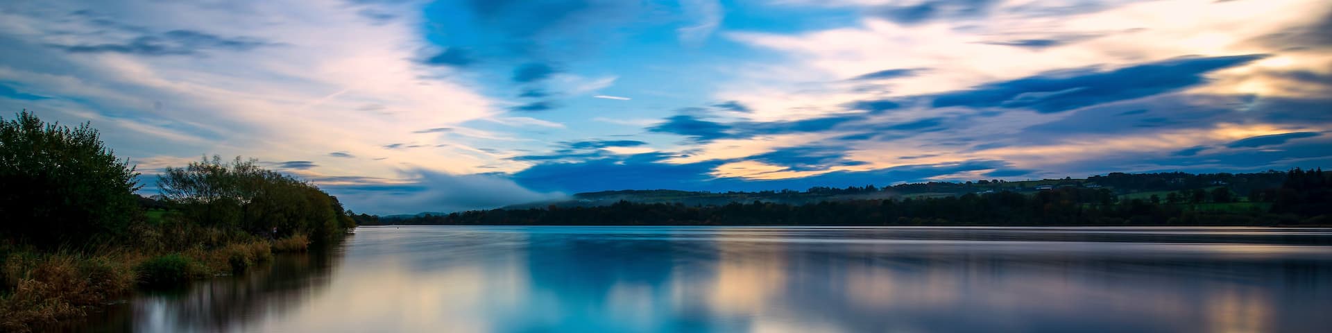 A calm river reflecting the sky, surrounded by trees and hills.