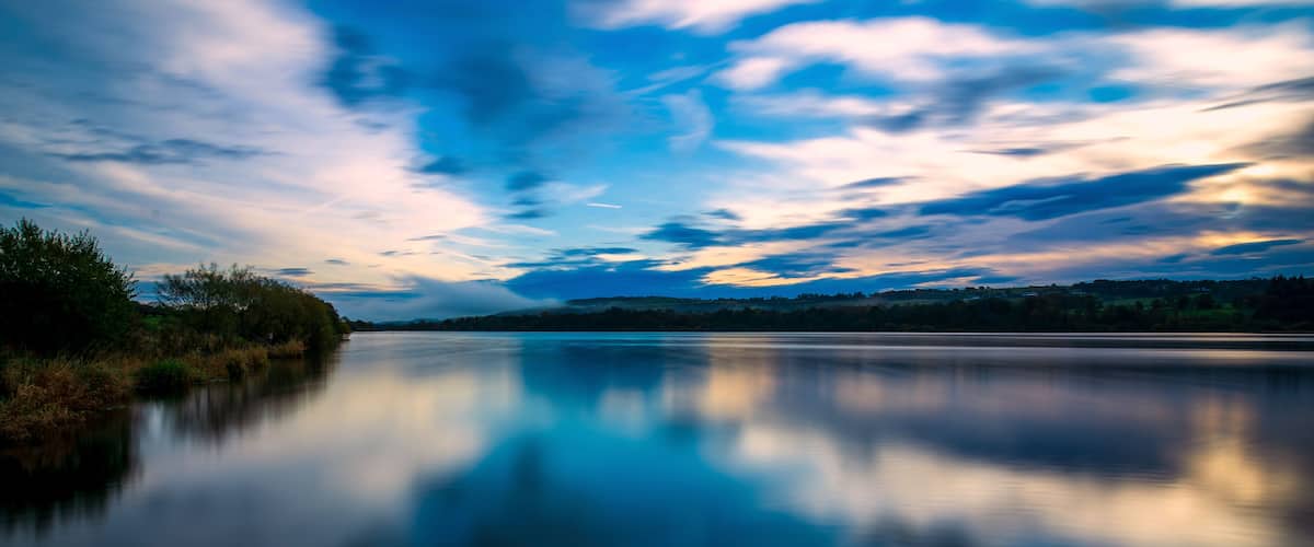 A calm river reflecting the sky, surrounded by trees and hills.