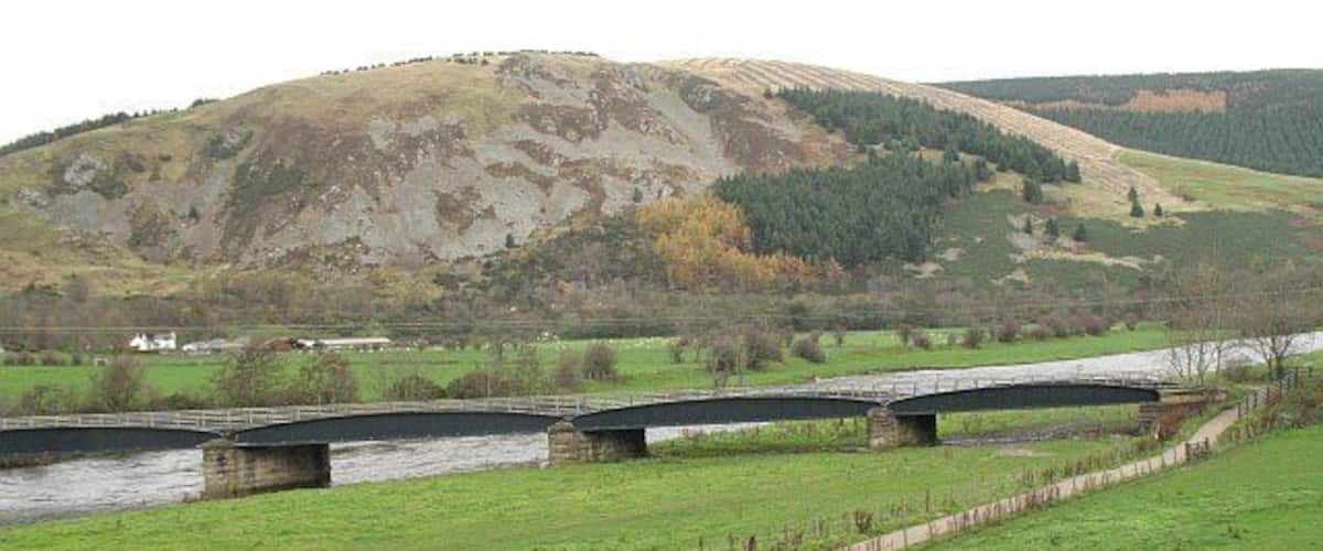 A river with three stone bridges, a grassy field, and a hill in the background.