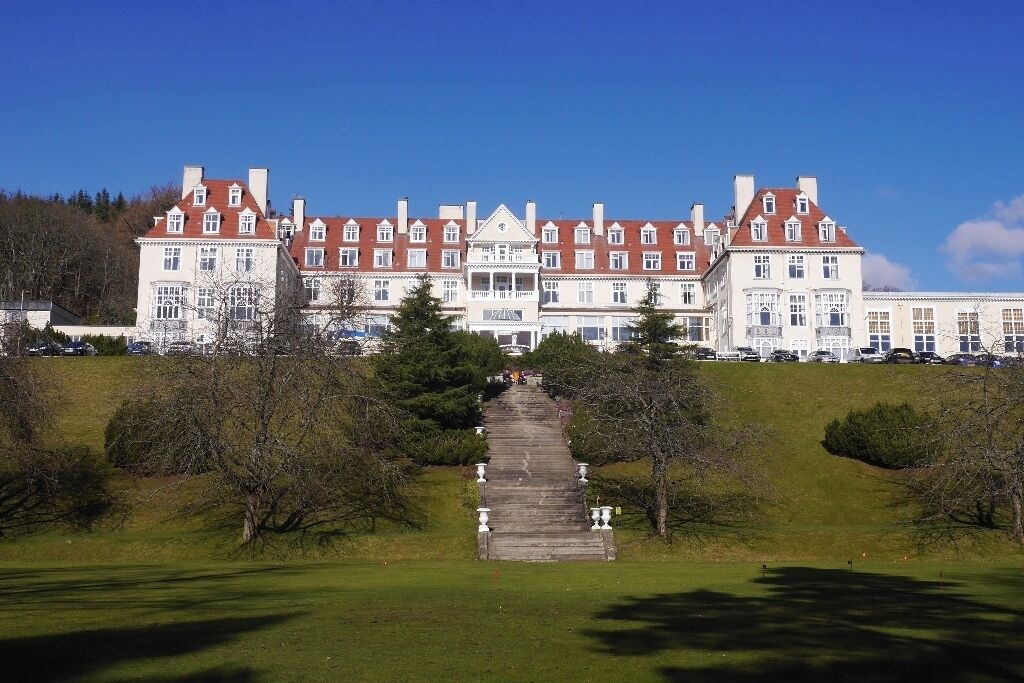A large, historic building with a symmetrical facade, a central entrance, and a surrounding lawn with trees.