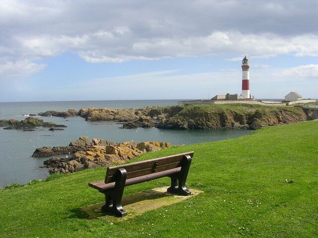 A lighthouse on a rocky outcrop, a grassy area with a bench, and the sea in the background.
