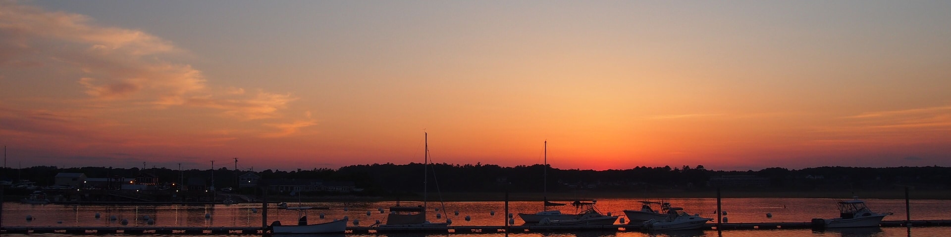 A sunset over a calm body of water with boats docked, a shoreline, and silhouettes of people.