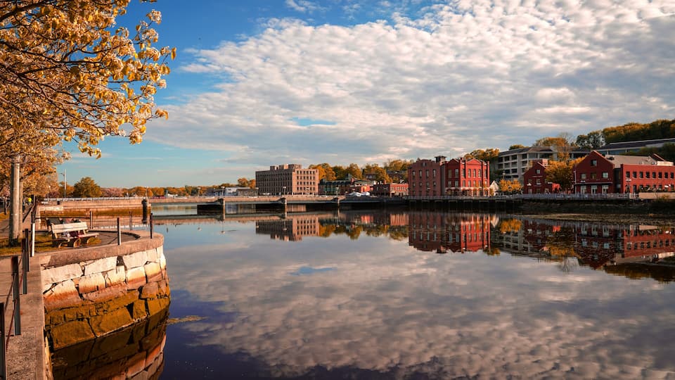A calm river reflecting buildings and trees, with a clear sky above.