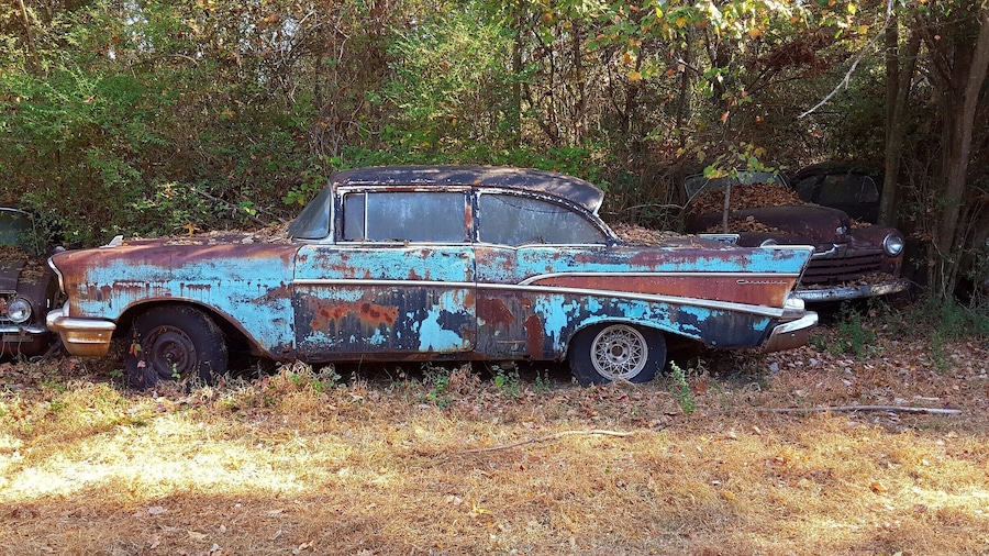 An old, rusted Chevrolet car in a wooded area.