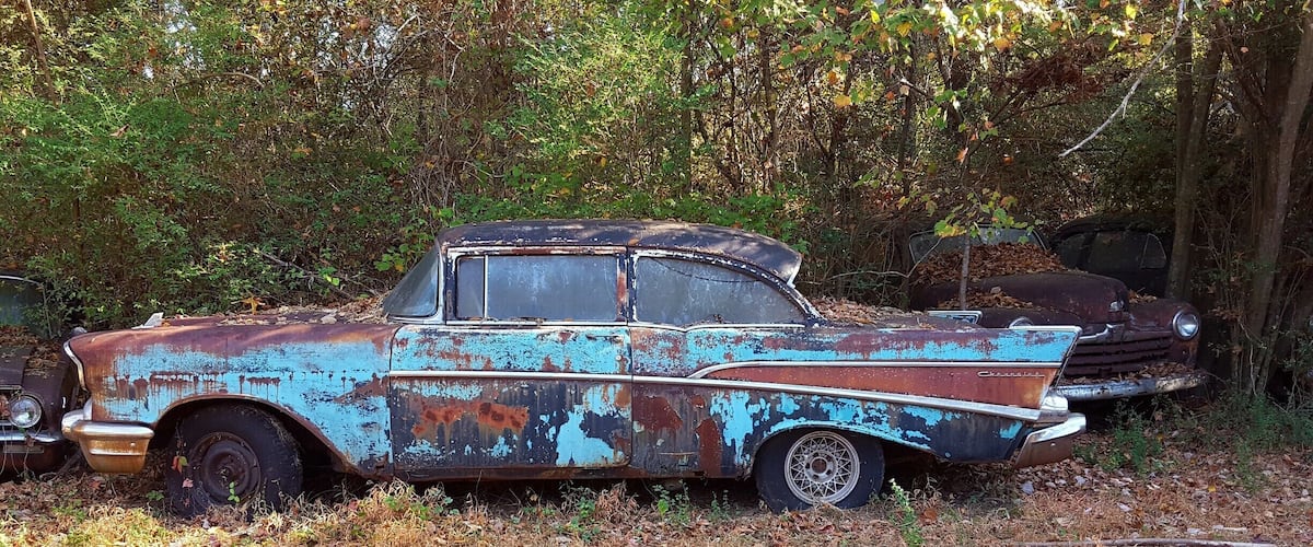 An old, rusted Chevrolet car in a wooded area.