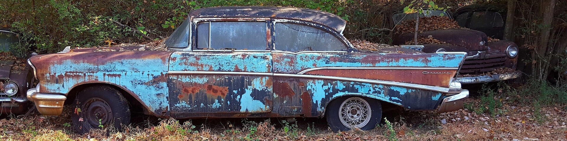 An old, rusted Chevrolet car in a wooded area.