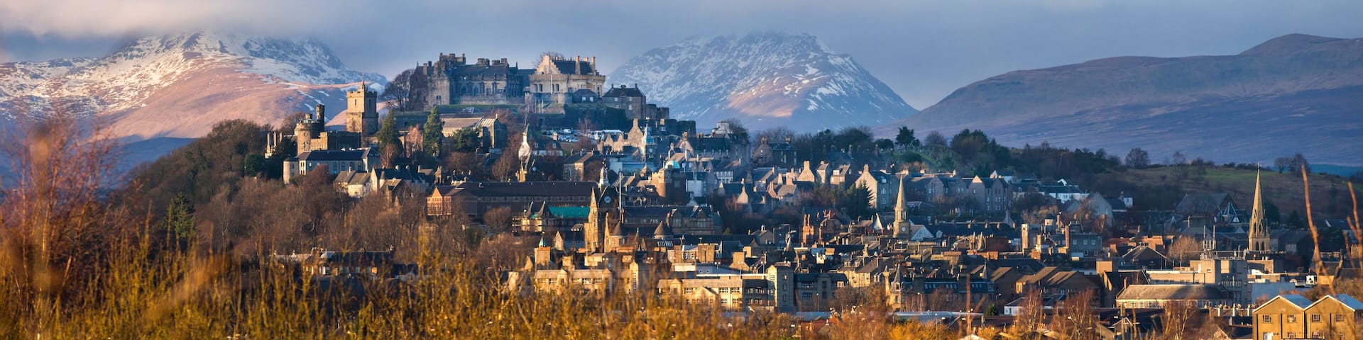 A historic town with medieval architecture, including a castle, set against a backdrop of mountains and a cloudy sky.