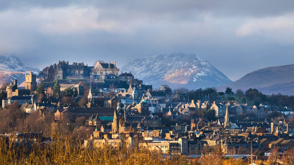 A historic town with medieval architecture, including a castle, set against a backdrop of mountains and a cloudy sky.