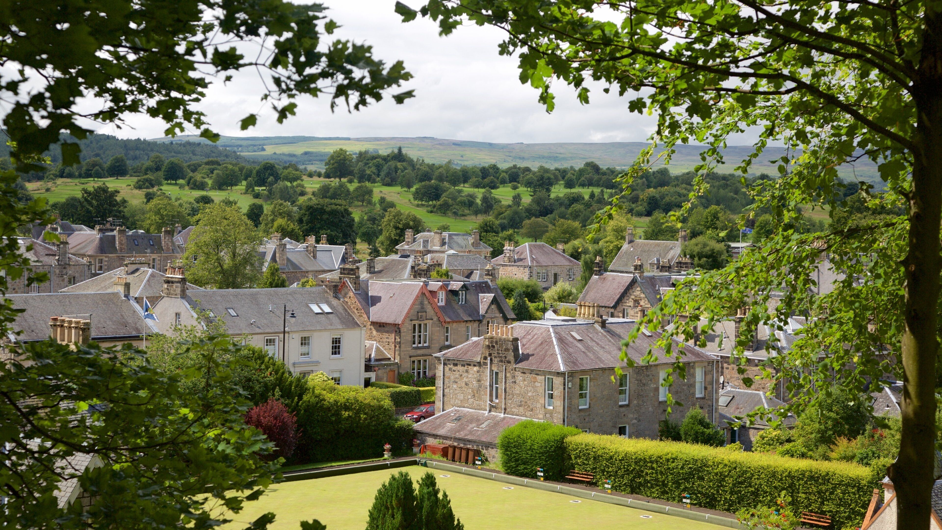 A village with stone houses, a green park, and rolling hills in the background.