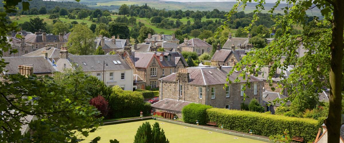 A village with stone houses, a green park, and rolling hills in the background.