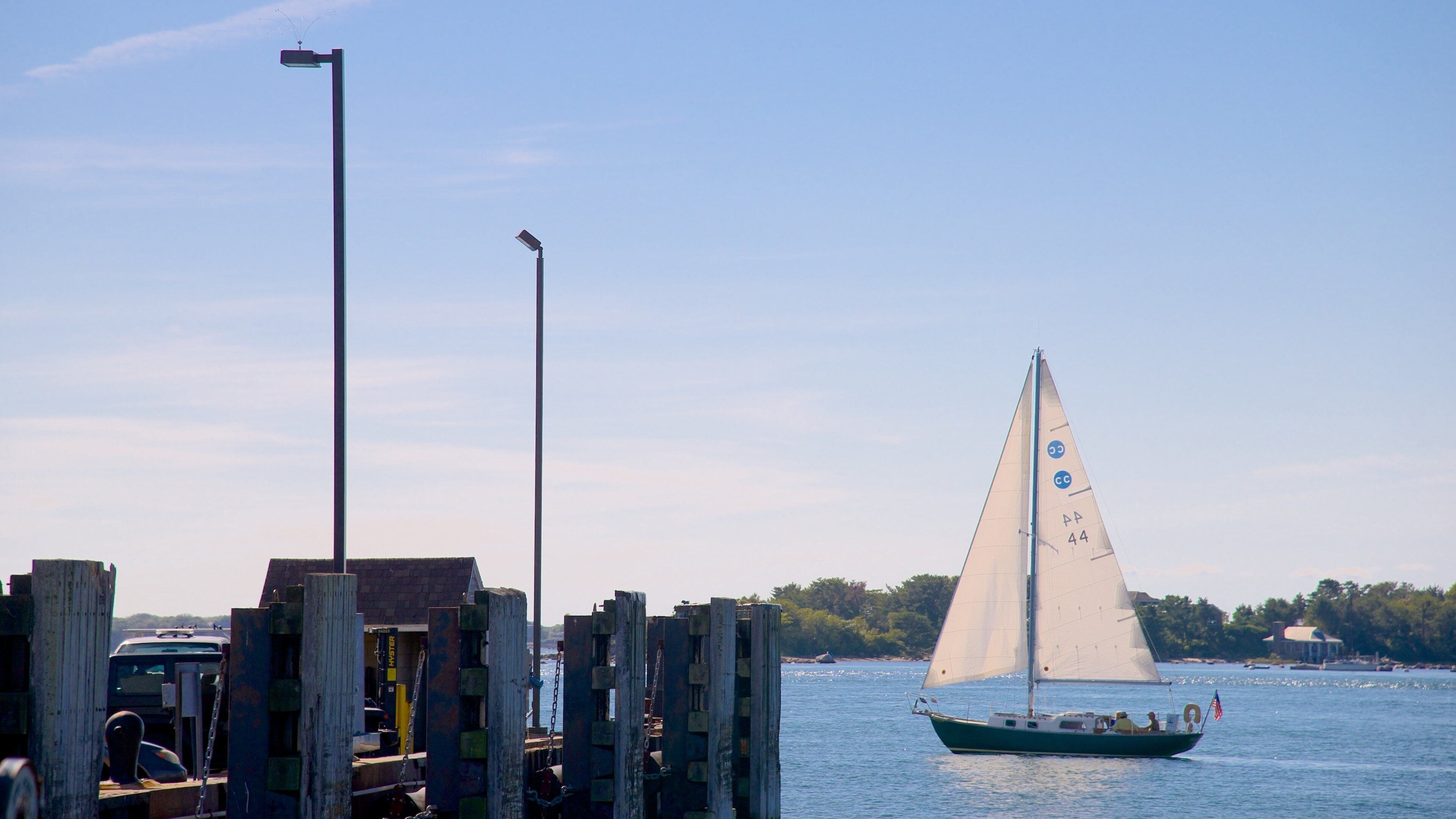 A sailboat on calm waters, docked at a wooden pier with a clear sky and distant trees.