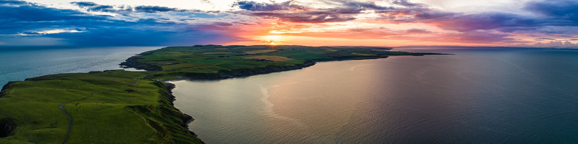 A coastal landscape with a winding road, a body of water, and a sunset.