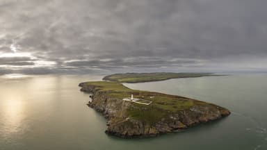 A coastal landscape with a lighthouse on a rocky outcrop, surrounded by calm waters and overcast skies.