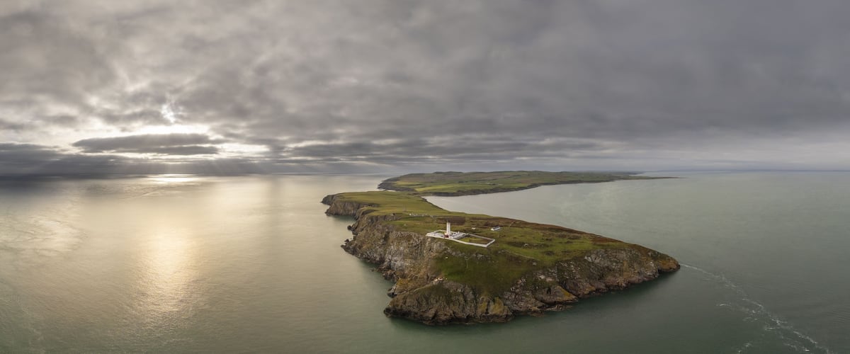 A coastal landscape with a lighthouse on a rocky outcrop, surrounded by calm waters and overcast skies.