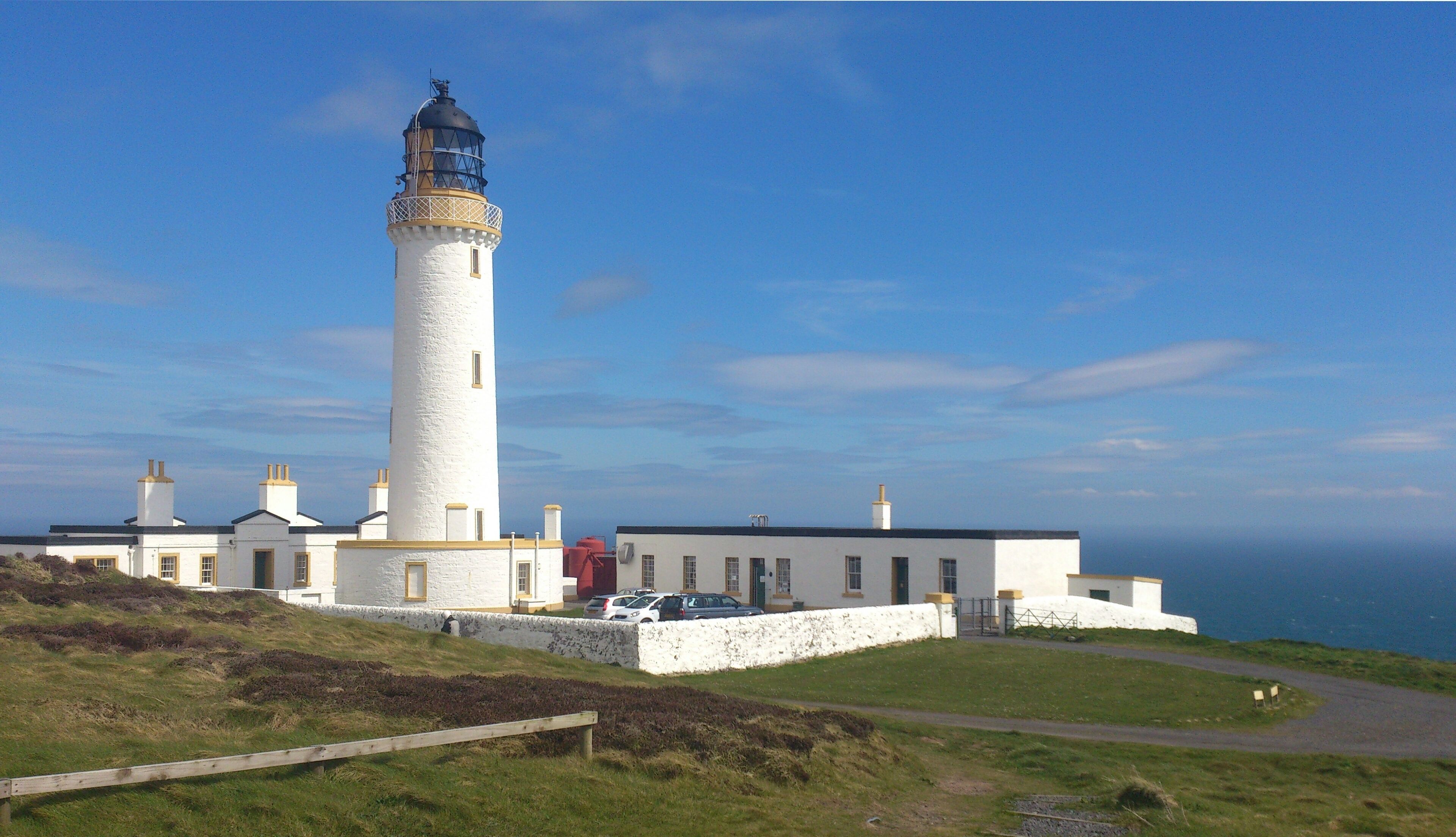 A lighthouse with a white tower and a black top, surrounded by a white building with multiple chimneys.