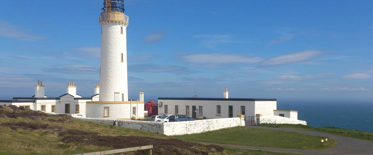 A lighthouse with a white tower and a black top, surrounded by a white building with multiple chimneys.