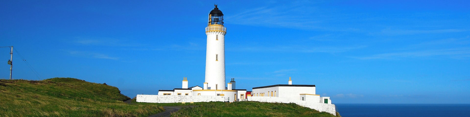 A white lighthouse with a black top, situated on a grassy hill with a clear blue sky and ocean in the background.