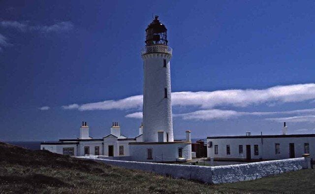 A tall white lighthouse with a black top, surrounded by a white building with multiple chimneys.