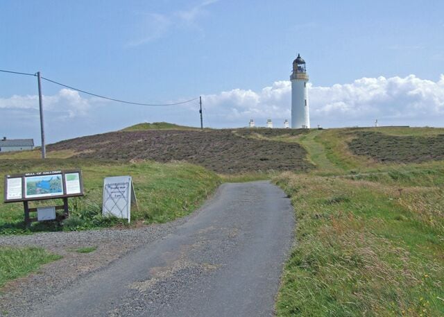 A coastal road leading to a lighthouse, with informational signs and a grassy hill in the background.