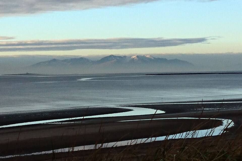 A coastal landscape with a sandy beach, a calm sea, and distant mountains.