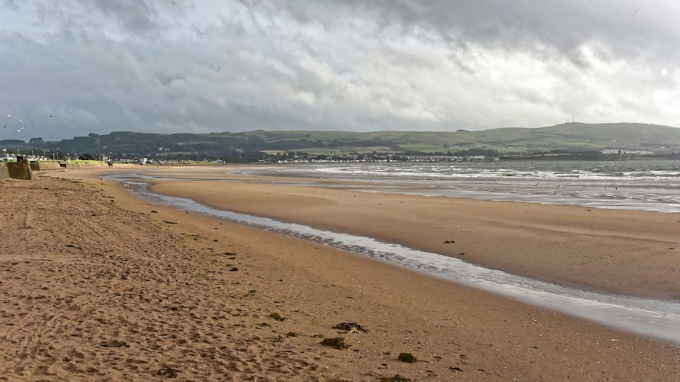 A sandy beach with gentle waves, a cloudy sky, and distant hills.
