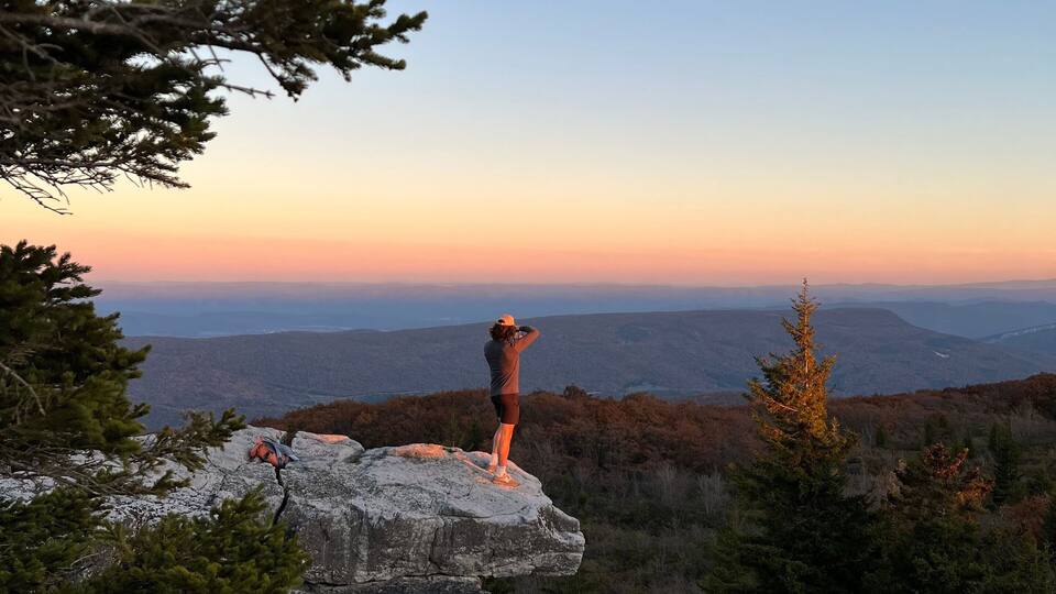 A person standing on a rocky outcrop overlooking a mountainous landscape during sunset.